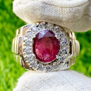 May include: A silver ring featuring a large oval ruby gemstone surrounded by smaller white diamonds. The ring has a textured band.