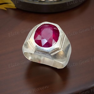 May include: A silver ring featuring a large, oval-cut ruby gemstone. The ruby is a deep red hue, set in a geometric bezel. The ring has a wide band and a polished finish. The ring is displayed on a dark brown surface.