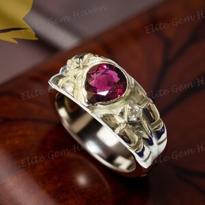 May include: A silver ring with a large, oval-cut, deep red gemstone as the centerpiece. The ring has a detailed, textured design with small accent stones. The ring is displayed on a dark, polished wooden surface.
