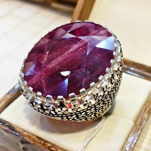 May include: A silver ring featuring a large, oval-shaped, faceted deep red gemstone set in a prong setting. The band of the ring is wide and decorated with a detailed, textured floral pattern that wraps around the entire circumference of the metal.