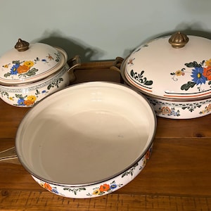 Puede incluir: Un juego de utensilios de cocina vintage de esmalte con diseños florales. El juego incluye dos ollas con tapa y una sartén. Las ollas y la sartén son blancas con flores coloridas y asas y perillas de color latón. Sobre una superficie de madera.