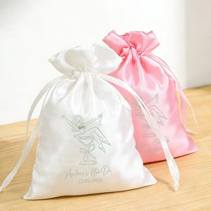 Puede incluir: Dos bolsas de satén con cordón, una blanca y otra rosa, sobre una superficie de madera. La bolsa blanca presenta una ilustración verde claro de una mujer en una copa de martini y el texto "Amber's Hen Do 22.05.2026". La bolsa rosa tiene un diseño similar.