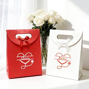 Puede incluir: Dos bolsas de regalo, una roja y otra blanca, con un diseño de corazón y estetoscopio. La bolsa roja tiene un lazo rojo y el nombre "Eliseo" en blanco. La bolsa blanca tiene un lazo blanco y el nombre "Maria" en rojo. Un ramo de rosas blancas está en el fondo.