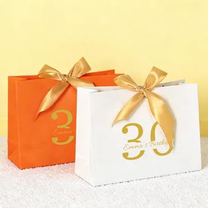 Könnte beinhalten: Zwei Geschenktüten, eine orangefarbene und eine weiße, mit goldenen Satinbändern. Die orangefarbene Tüte hat eine goldene "30" und den Text "Emma's Birthday". Die weiße Tüte hat eine goldene "30" und den Text "Emma's Birthday". Die Tüten stehen auf einer weißen Oberfläche.