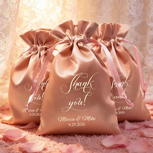 Op de afbeelding: Drie blush roze satijnen trekkoordzakjes met de woorden "Thank you!" in sierlijk wit schrift. De zakjes bevatten ook de namen "Maria & Mike" en de datum "8.29.2026". Roze linten maken het ontwerp compleet.