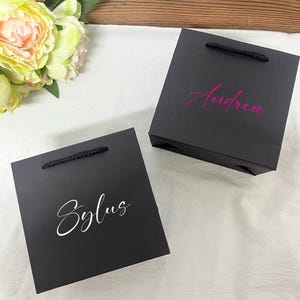 Peut inclure: Deux sacs cadeaux noirs avec des poignées en corde. L'un porte le nom "Sylus" en écriture blanche, l'autre le nom "Andrea" en écriture rose. Les sacs sont sur une surface blanche avec un arrangement floral et un fond en bois.