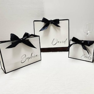 Peut inclure: Trois sacs cadeaux blancs avec une bordure noire et des n&oelig;uds en satin noir. Les sacs portent des noms &eacute;crits en script &eacute;l&eacute;gant : Sophia, David et Emily. Les sacs sont dispos&eacute;s sur une surface blanche.