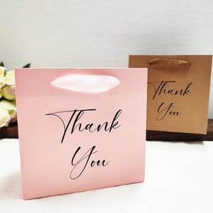 May include: Two small gift bags. One is light pink with a white handle and the words "Thank You" in black script. The other is brown with a brown handle and the same text. Both bags are square.