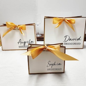 Peut inclure: Trois sacs cadeaux blancs avec des bordures et des poign&eacute;es dor&eacute;es, chacun orn&eacute; d'un n&oelig;ud en satin dor&eacute;. Les sacs sont personnalis&eacute;s avec des noms et le mot "Bridesmaid" en &eacute;criture noire. Un verre &agrave; vin est en arri&egrave;re-plan.