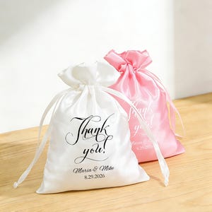 May include: Two satin drawstring bags, one white and one pink, are displayed on a wooden surface. The white bag features the text "Thank you!" in script, plus a name and date. The pink bag has a similar design. Both bags are tied with satin ribbons.