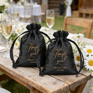 May include: Two black satin drawstring favour bags with gold text reading "Thank you!" and the names "Maria & Mike" with a date. The bags are on a wooden table with champagne flutes and flowers.