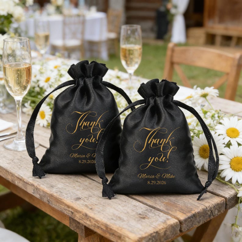 May include: Two black satin drawstring favour bags with gold text reading "Thank you!" and the names "Maria & Mike" with a date. The bags are on a wooden table with champagne flutes and flowers.