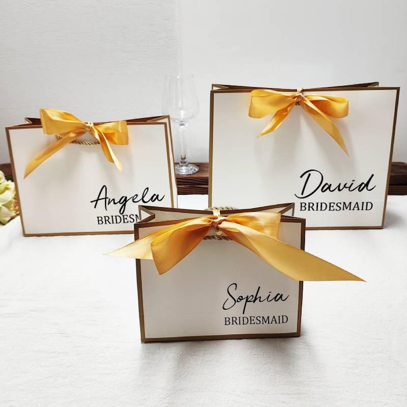 Peut inclure: Trois sacs cadeaux blancs avec des bordures et des poign&eacute;es dor&eacute;es, chacun orn&eacute; d'un n&oelig;ud en satin dor&eacute;. Les sacs sont personnalis&eacute;s avec des noms et le mot "Bridesmaid" en &eacute;criture noire. Un verre &agrave; vin est en arri&egrave;re-plan.