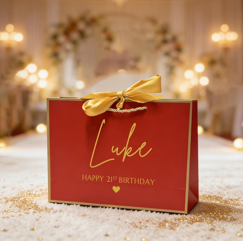 Peut inclure: Un sac cadeau rouge avec une bordure dor&eacute;e et un n&oelig;ud dor&eacute;. Le sac porte le nom "Luke" en lettres dor&eacute;es, ainsi que "HAPPY 21ST BIRTHDAY" et un c&oelig;ur dor&eacute;. Le sac est pos&eacute; sur une surface blanche.