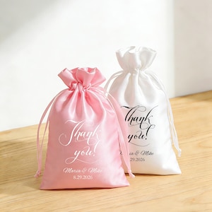 May include: Two satin drawstring bags, one pink and one white, with "Thank you!" printed in elegant script. The pink bag also includes "Maria & Mike 8.29.2026". Both bags are tied at the top with matching satin cords.