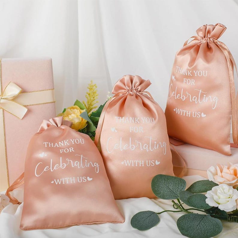 May include: Peach satin drawstring bags with the text "THANK YOU FOR Celebrating WITH US" printed in white. The bags are gathered at the top with matching satin cords. A wrapped gift box and flowers are in the background.