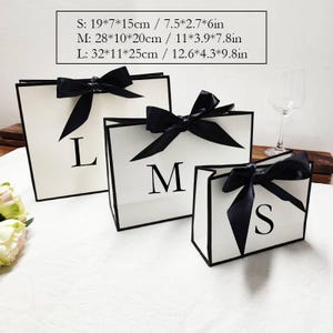 Peut inclure: Trois sacs cadeaux blancs avec bordure et n&oelig;uds noirs. Les sacs sont &eacute;tiquet&eacute;s avec les lettres L, M et S. Les dimensions sont fournies en centim&egrave;tres et en pouces. Le plus grand sac mesure 32 x 11 x 25 cm.