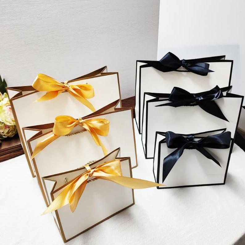 Peut inclure: Sacs cadeaux de diff&eacute;rentes tailles. Les sacs sont blancs avec des bordures dor&eacute;es et des n&oelig;uds en ruban de satin dor&eacute;. D'autres sacs sont blancs avec des bordures noires et des n&oelig;uds en ruban de satin noir. Sur une surface blanche.