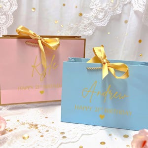 Peut inclure: Deux sacs cadeaux, un rose et un bleu, avec des d&eacute;tails dor&eacute;s. Le sac rose porte le nom "Kyle" et le sac bleu porte le nom "Andrew" en lettres dor&eacute;es. Les deux sacs indiquent "Happy 21st Birthday" avec un c&oelig;ur dor&eacute;. Chaque sac a un ruban dor&eacute;.