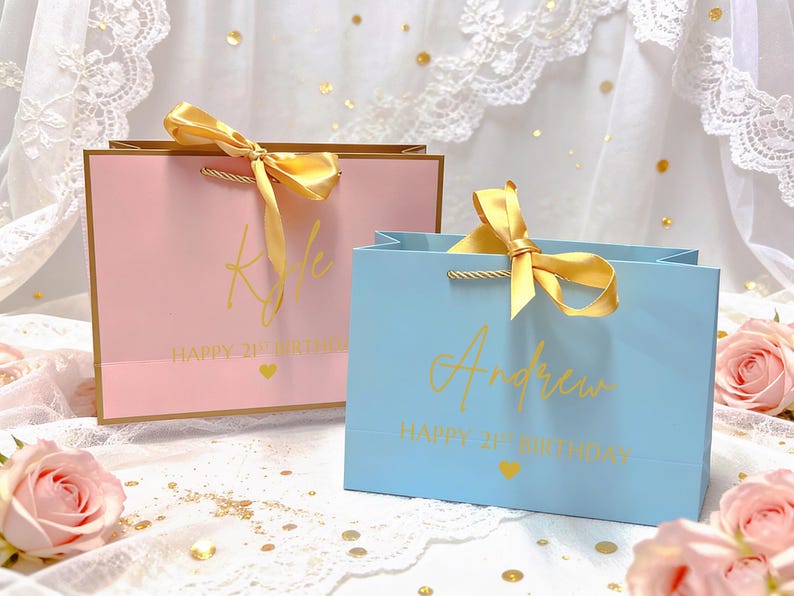 Peut inclure: Deux sacs cadeaux, un rose et un bleu, avec des d&eacute;tails dor&eacute;s. Le sac rose porte le nom "Kyle" et le sac bleu porte le nom "Andrew" en lettres dor&eacute;es. Les deux sacs indiquent "Happy 21st Birthday" avec un c&oelig;ur dor&eacute;. Chaque sac a un ruban dor&eacute;.
