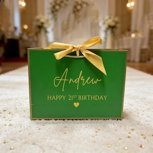 Peut inclure: Un sac cadeau vert avec des bordures dor&eacute;es et un ruban dor&eacute;. Le sac porte le nom "Andrew" et le texte "HAPPY 21ST BIRTHDAY" en lettres dor&eacute;es, ainsi qu'un c&oelig;ur dor&eacute;. Le sac est pos&eacute; sur un tapis blanc.