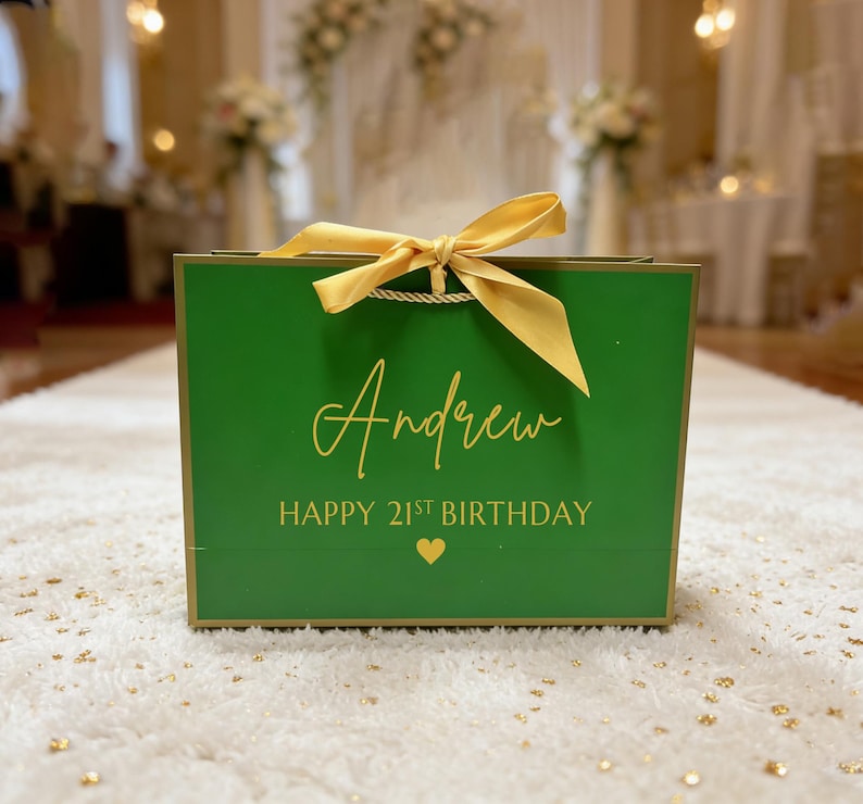 Peut inclure: Un sac cadeau vert avec des bordures dor&eacute;es et un ruban dor&eacute;. Le sac porte le nom "Andrew" et le texte "HAPPY 21ST BIRTHDAY" en lettres dor&eacute;es, ainsi qu'un c&oelig;ur dor&eacute;. Le sac est pos&eacute; sur un tapis blanc.