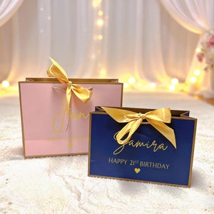 Peut inclure: Deux sacs cadeaux, un rose et un bleu marine, avec des bordures et des n&oelig;uds dor&eacute;s. Le sac rose porte le nom "Jon" en &eacute;criture dor&eacute;e, et le sac bleu porte le nom "Samira". Les deux sacs indiquent "Happy 21st Birthday" en or.