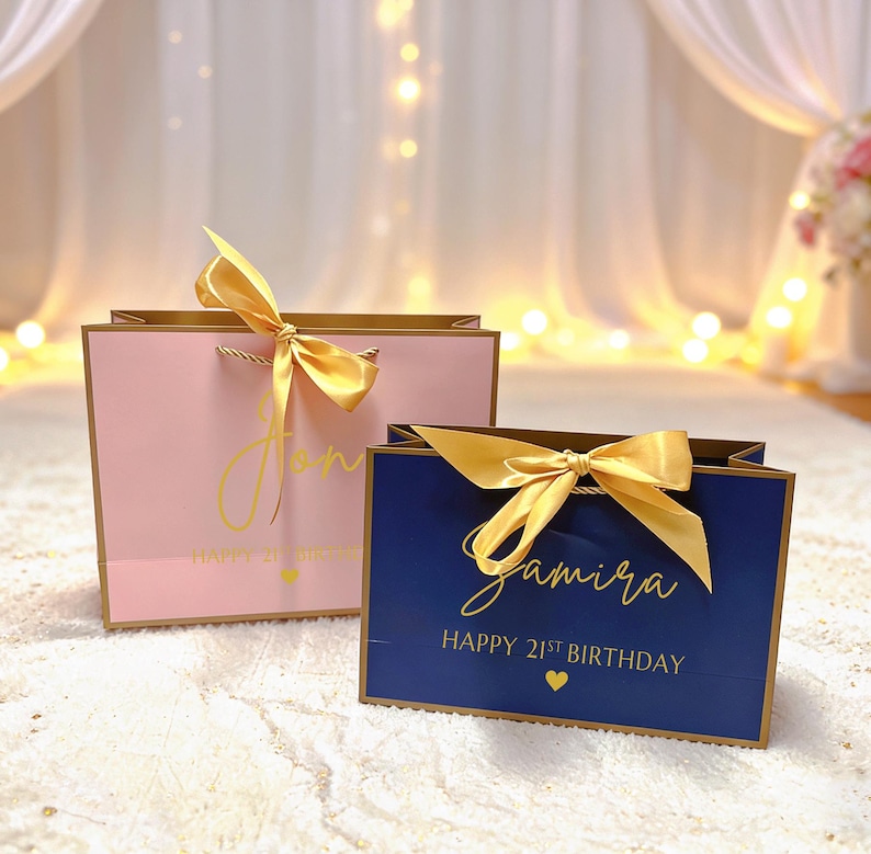 Peut inclure: Deux sacs cadeaux, un rose et un bleu marine, avec des bordures et des n&oelig;uds dor&eacute;s. Le sac rose porte le nom "Jon" en &eacute;criture dor&eacute;e, et le sac bleu porte le nom "Samira". Les deux sacs indiquent "Happy 21st Birthday" en or.