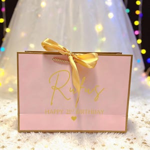 Peut inclure: Un sac cadeau rose avec une bordure dor&eacute;e et un n&oelig;ud en satin dor&eacute;. Le sac porte le nom "Rufus" en lettres dor&eacute;es, ainsi que "HAPPY 21ST BIRTHDAY" et un c&oelig;ur dor&eacute;.