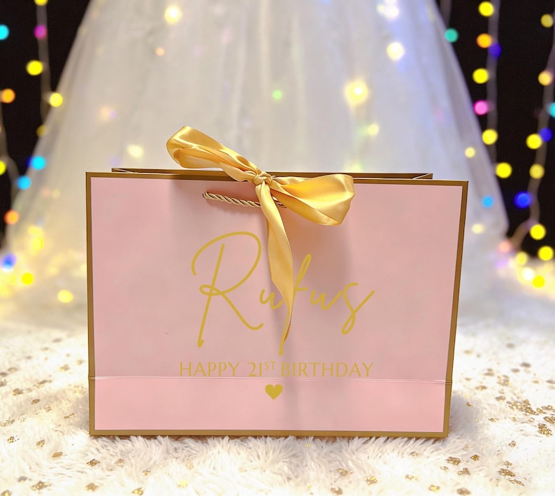 Peut inclure: Un sac cadeau rose avec une bordure dor&eacute;e et un n&oelig;ud en satin dor&eacute;. Le sac porte le nom "Rufus" en lettres dor&eacute;es, ainsi que "HAPPY 21ST BIRTHDAY" et un c&oelig;ur dor&eacute;.