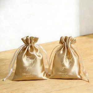 May include: Two gold satin drawstring bags with white text reading "Thank you!" and the names "Maria & Mike" with the date "8.29.2026". The bags are tied with white satin ribbons.