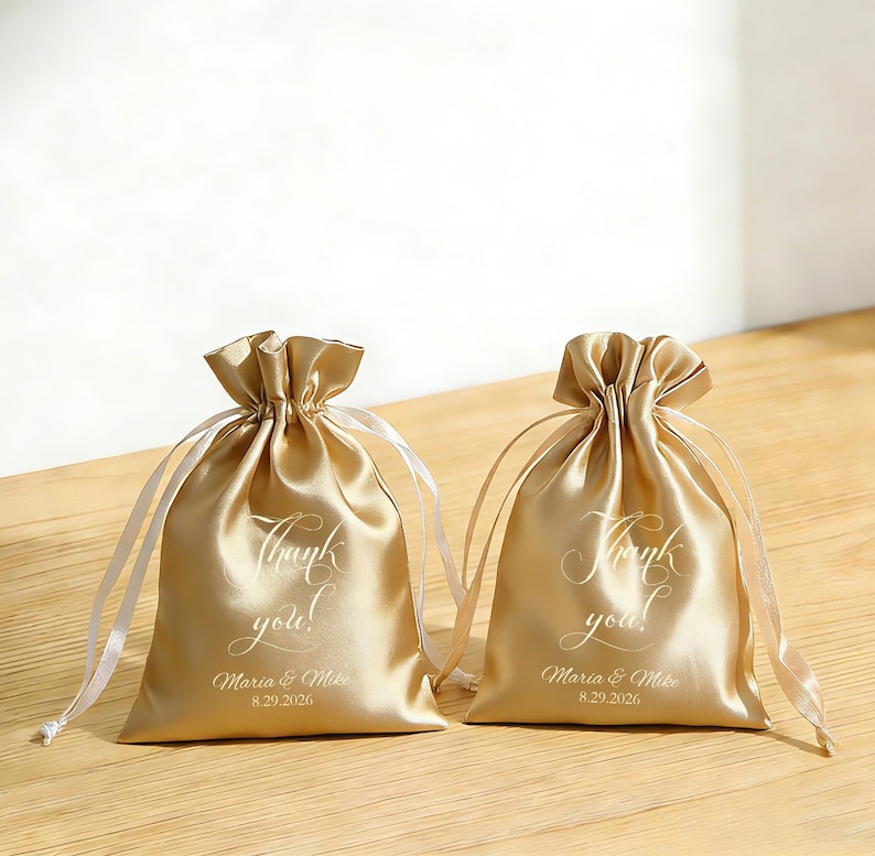 May include: Two gold satin drawstring bags with white text reading "Thank you!" and the names "Maria & Mike" with the date "8.29.2026". The bags are tied with white satin ribbons.