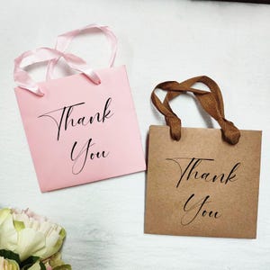 May include: Two small gift bags, one pink and one brown, with the words "Thank You" in elegant script. The pink bag has satin ribbon handles, while the brown bag has woven brown handles. Ideal for small gifts.