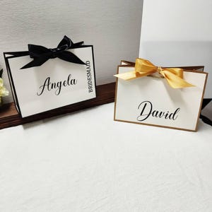 Peut inclure: Deux sacs cadeaux avec des fonds blancs et des bordures noires. L'un des sacs a un n&oelig;ud noir et le nom "Angela" en cursive, avec "Bridesmaid" imprim&eacute; verticalement. L'autre sac a un n&oelig;ud dor&eacute; et le nom "David" en cursive.