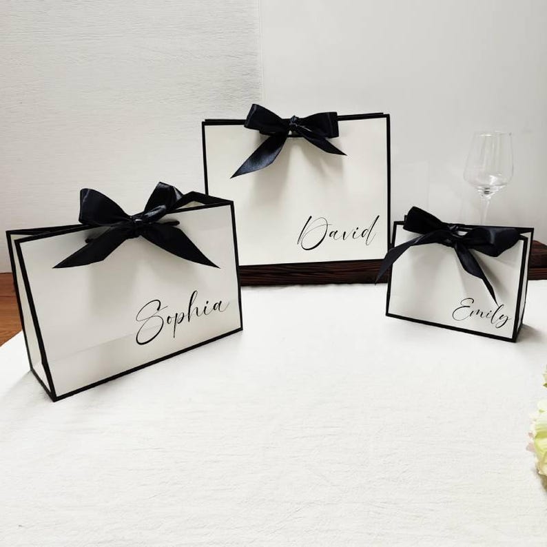 Peut inclure: Trois sacs cadeaux blancs avec une bordure noire et des n&oelig;uds en satin noir. Les sacs portent des noms &eacute;crits en script &eacute;l&eacute;gant : Sophia, David et Emily. Les sacs sont dispos&eacute;s sur une surface blanche.