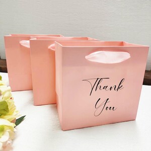 May include: Three pale pink gift bags with white handles. The front bag has the words "Thank You" in black script. The bags are square and have a glossy finish, ideal for small gifts or presents.