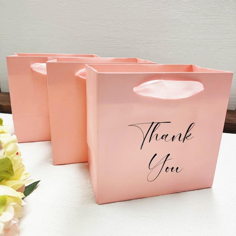 May include: Three pale pink gift bags with white handles. The front bag has the words "Thank You" in black script. The bags are square and have a glossy finish, ideal for small gifts or presents.