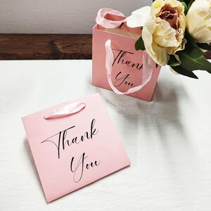 May include: Two pink gift bags, each with the words "Thank You" in black script. One bag lies flat, the other stands with a white ribbon handle. A bouquet of cream-coloured flowers is in the background.