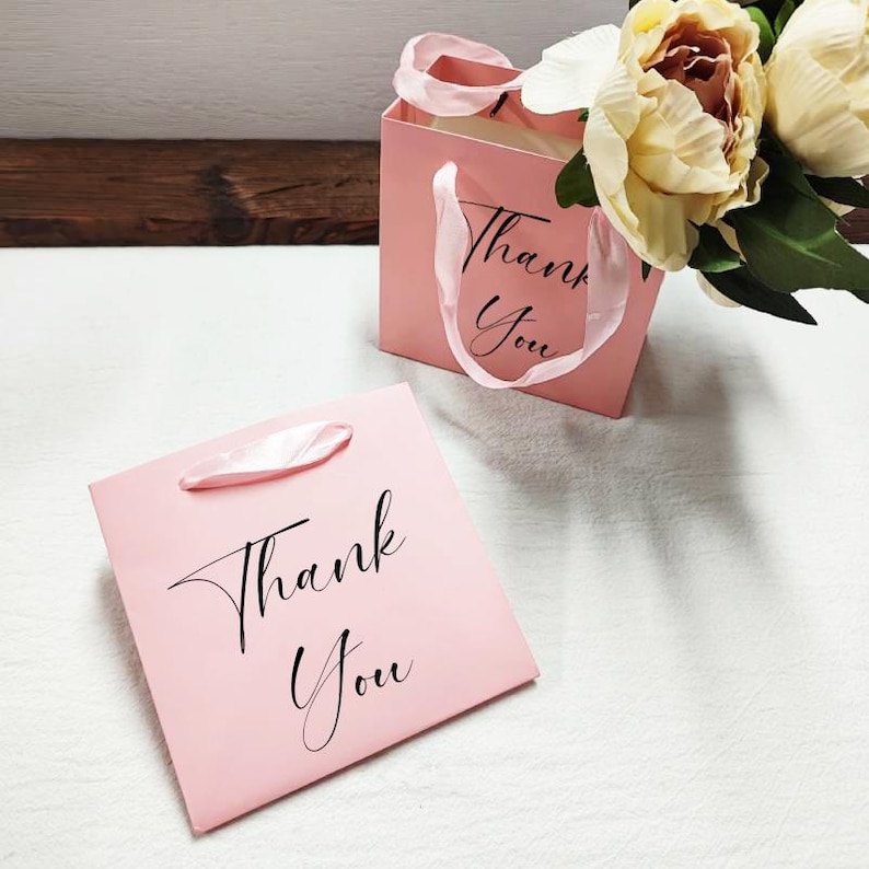 May include: Two pink gift bags, each with the words "Thank You" in black script. One bag lies flat, the other stands with a white ribbon handle. A bouquet of cream-coloured flowers is in the background.