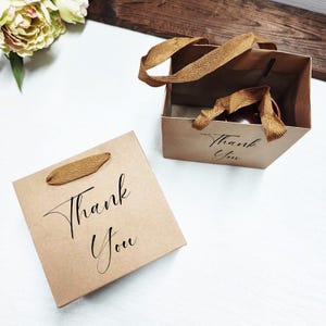 May include: Two small, brown paper gift bags with woven handles. One bag is open, and the other is closed. Both bags have the words "Thank You" printed in black script. The bags are on a white surface.