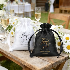 May include: Two satin drawstring favour bags, one white and one black, sit on a wooden table. The white bag has black text that says "Thank you!" and the date. The black bag has gold text that says "Thank you!" and the date.