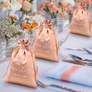 May include: Peach satin drawstring favour bags with white text reading "Thank You for Celebrating with Us." The bags are tied with matching satin ribbon. Silverware and a wine glass are on a white tablecloth, with floral arrangements in the background.