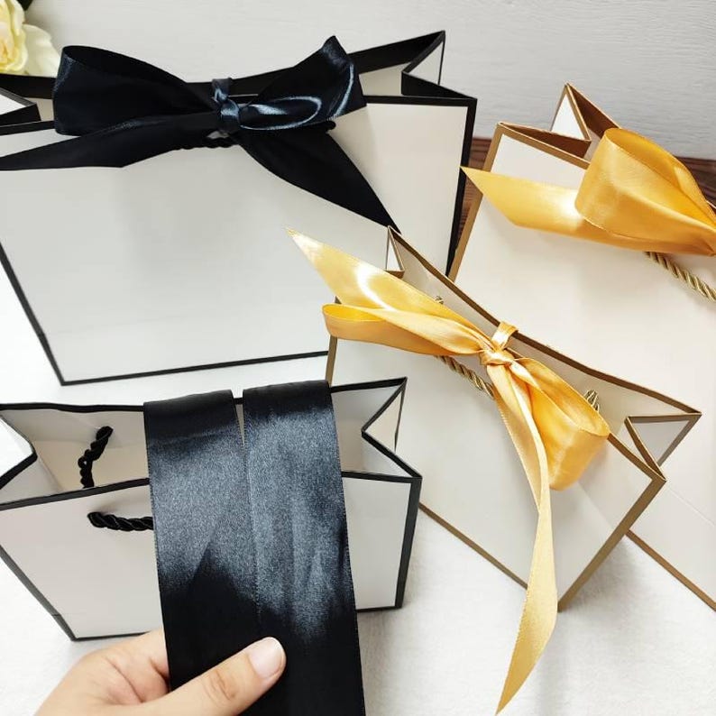 Peut inclure: Sacs cadeaux blancs avec bordures et poign&eacute;es noires, orn&eacute;s de n&oelig;uds en satin noir. Un sac est orn&eacute; d'un n&oelig;ud en satin dor&eacute;. Les sacs sont con&ccedil;us pour offrir et ont une apparence luxueuse.