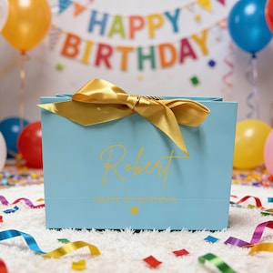 Peut inclure: Un sac cadeau bleu clair avec un n&oelig;ud dor&eacute; et le nom "Robert" en lettres dor&eacute;es. Le sac porte l'inscription "HAPPY 1st BIRTHDAY" et un petit c&oelig;ur dor&eacute;. Des ballons et une banni&egrave;re "HAPPY BIRTHDAY" sont en arri&egrave;re-plan.