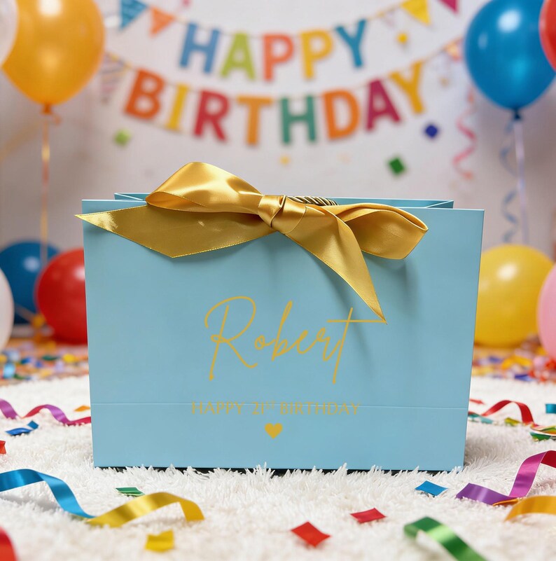Peut inclure: Un sac cadeau bleu clair avec un n&oelig;ud dor&eacute; et le nom "Robert" en lettres dor&eacute;es. Le sac porte l'inscription "HAPPY 1st BIRTHDAY" et un petit c&oelig;ur dor&eacute;. Des ballons et une banni&egrave;re "HAPPY BIRTHDAY" sont en arri&egrave;re-plan.