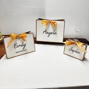 Peut inclure: Trois sacs cadeaux blancs avec des bordures et des n&oelig;uds dor&eacute;s. Chaque sac porte un nom &eacute;crit en script noir : "Emily" avec "Bridesmaid" en dessous, "Angela" et "Sophia". Les sacs sont dispos&eacute;s sur une surface blanche.