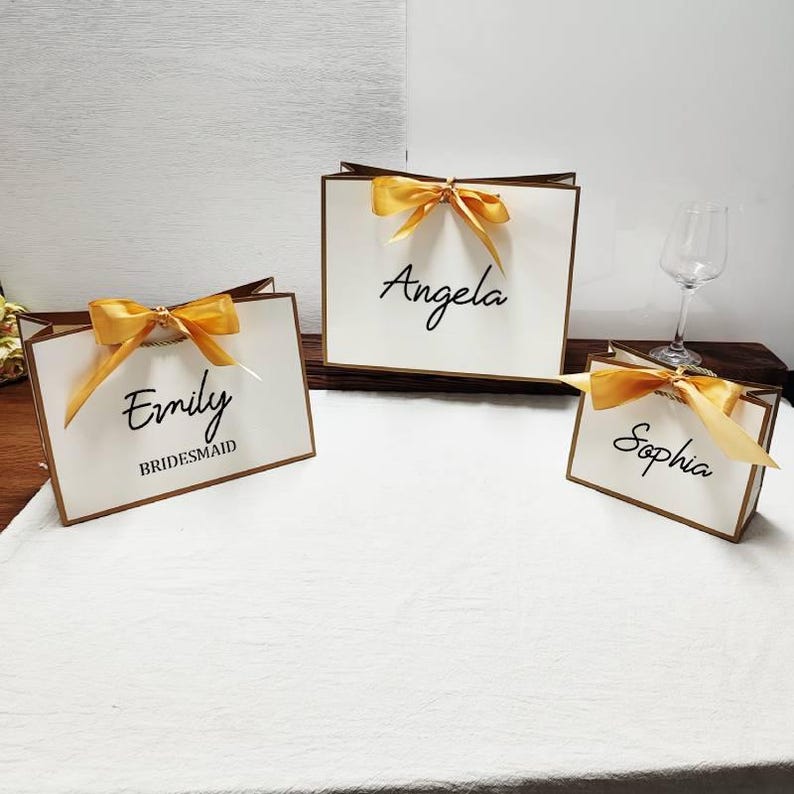 Peut inclure: Trois sacs cadeaux blancs avec des bordures et des n&oelig;uds dor&eacute;s. Chaque sac porte un nom &eacute;crit en script noir : "Emily" avec "Bridesmaid" en dessous, "Angela" et "Sophia". Les sacs sont dispos&eacute;s sur une surface blanche.