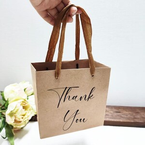May include: A small, brown paper gift bag with the words "Thank You" in elegant black script. The bag has brown ribbon handles and is held up by a hand. The background includes a bouquet of white and yellow flowers.