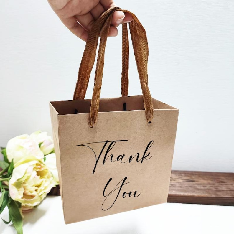 May include: A small, brown paper gift bag with the words "Thank You" in elegant black script. The bag has brown ribbon handles and is held up by a hand. The background includes a bouquet of white and yellow flowers.