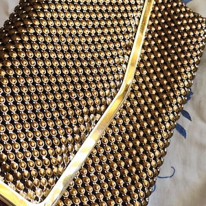 May include: A rectangular clutch bag featuring a metallic bronze mesh exterior composed of small, uniform circular studs. The bag is finished with a smooth, polished gold-tone metal frame along the top edge, providing a structured closure.